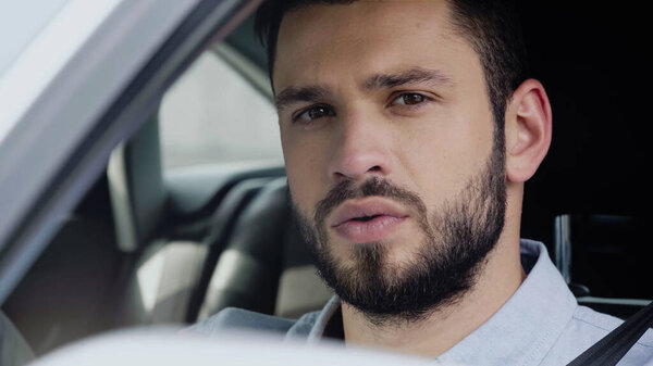 portrait of young and bearded man looking at camera while traveling in car on blurred foreground
