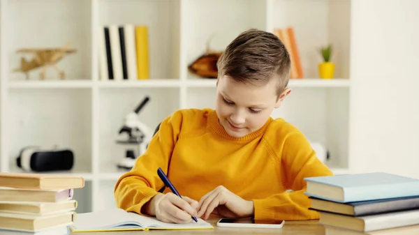 cheerful schoolboy writing in copy book near smartphone and books on desk