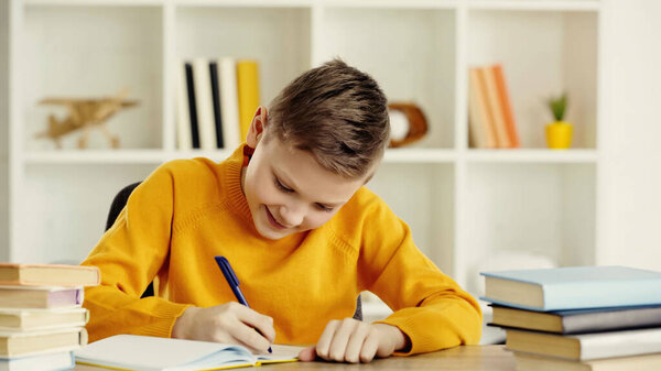 smiling preteen boy writing in notebook while doing homework near books on wooden table at home