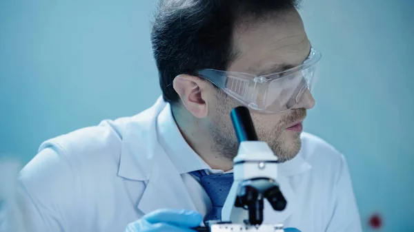 handsome scientist in goggles looking away near microscope in laboratory