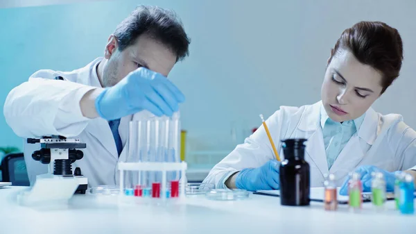 scientist taking test tube with sample near colleague writing in laboratory