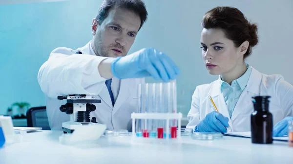 scientist taking test tube with sample near colleague with pencil in lab