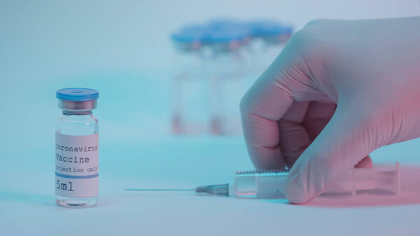 cropped view of person taking syringe near bottles with coronavirus vaccine on blue