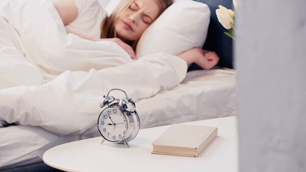 Blurred woman sleeping on bed near alarm clock and book in bedroom 