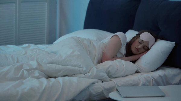 Woman in mask sleeping on bed near laptop on bedside table 