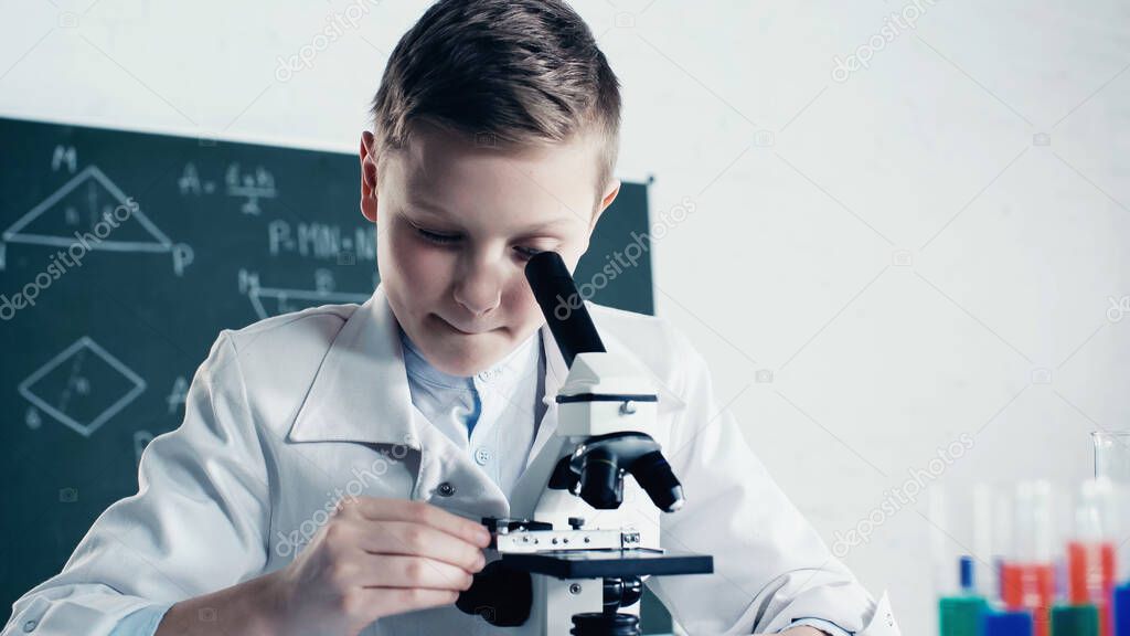 schoolkid in white coat looking through microscope during chemistry lesson in classroom  