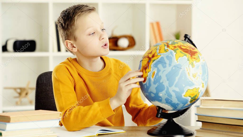 schoolboy in yellow jumper looking at globe near notebook and books on desk 