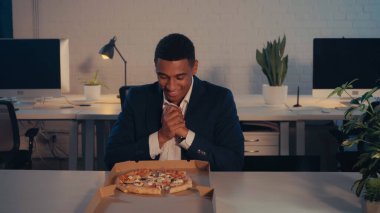 Gülümseyen Afro-Amerikan iş adamı geceleri ofiste pizzaya bakıyor.