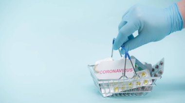 Plastik eldiven giymiş, elinde Coronavirus harfleriyle alışveriş sepeti tutan ve mavi üzerine ilaç koyan birinin kırpılmış görüntüsü.  