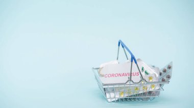 Üzerinde koronavirüs harfleri olan küçük bir alışveriş sepeti ve mavi üzerine ilaçlar. 
