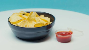 Çıtır çıtır patates cipsleri plastik kabın yanında, üzerinde mavi ketçap var.