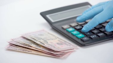 Beyaz arkaplandaki dolarların yakınındaki hesap makinesini kullanan lateks eldivenli kişinin kırpılmış görüntüsü