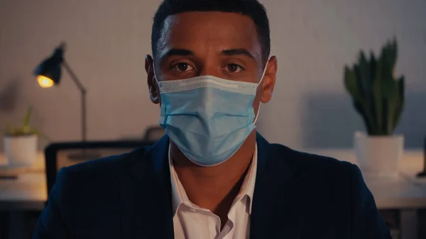 African american businessman in medical mask looking at camera in office at night