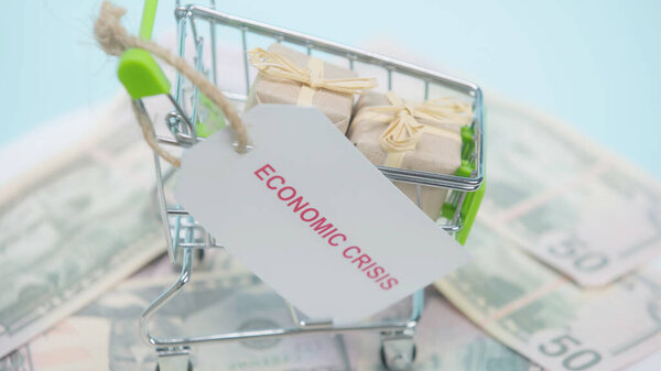 close up of shopping cart with gifts and card with economic crisis lettering on dollars and blue background 