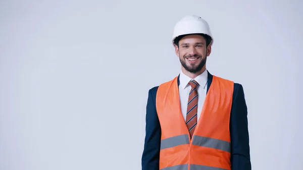 smiling engineer in hardhat and safety vest looking at camera isolated on grey