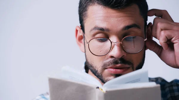 thoughtful nerd student in eyeglasses reading blurred book and touching head isolated on grey