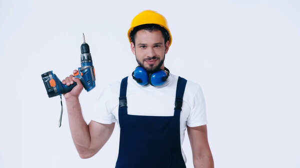 positive builder in hardhat and overalls holding electric drill and looking at camera isolated on white