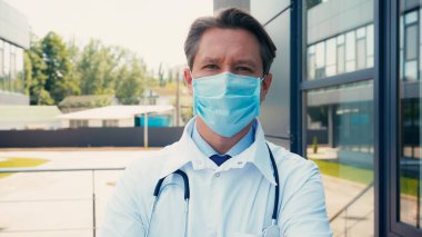 Tıbbi maskeli ve steteskoplu beyaz önlüklü doktor dışarıdaki kameraya bakıyor.