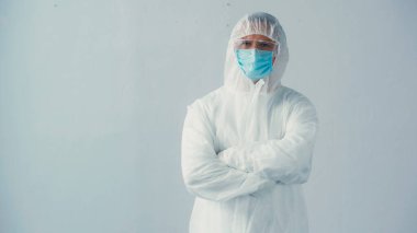Koruyucu giysili doktor ve gözlüklü tıbbi maske gri arka planda çapraz kollarla ayakta duruyor.