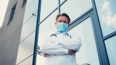 Doktor maskesinin düşük açılı görüntüsü kollarını kavuşturup kameraya bakıyor.