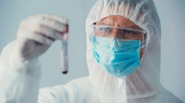 Tehlikeli madde giysisi ve gözlüklü virolog gri renkli test tüpüne bakıyor.