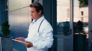 Stetoskop ve dijital tabletli doktor hastane binasının yanında duruyor.