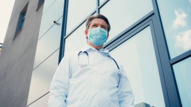 Klinik binasının yakınında beyaz önlüklü ve tıbbi maskeli doktorun düşük açılı görüntüsü