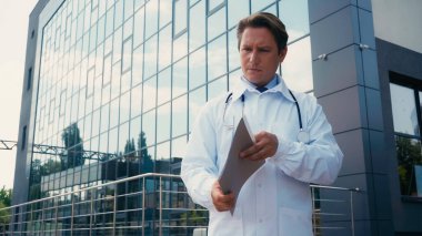 Beyaz önlüklü ciddi bir doktor hastanenin yanında cam cepheli belgeler tutuyor.