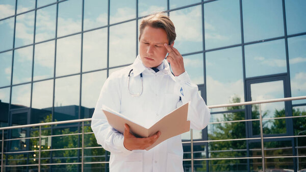 вдумчивый доктор трогает голову, глядя в папку с документами на открытом воздухе