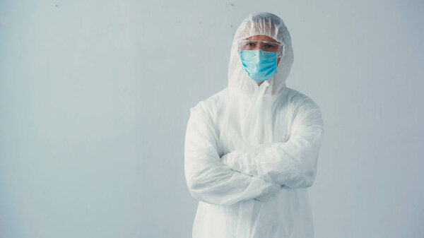 doctor in hazmat suit and medical mask with goggles standing with crossed arms on grey background