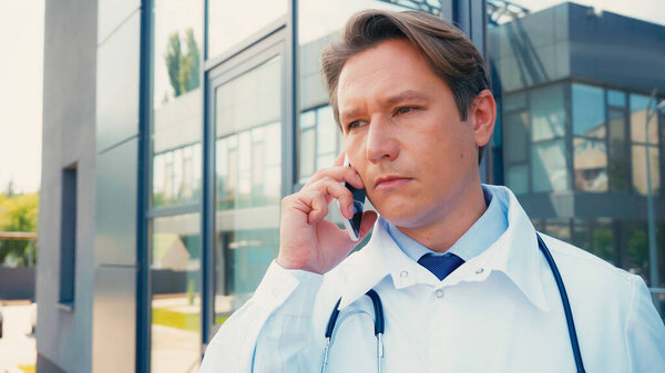 серьезный врач разговаривает по мобильному телефону возле городской больницы на открытом воздухе