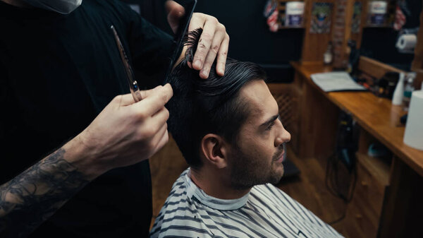 Tattooed hairdresser combing hair of client in barbershop 
