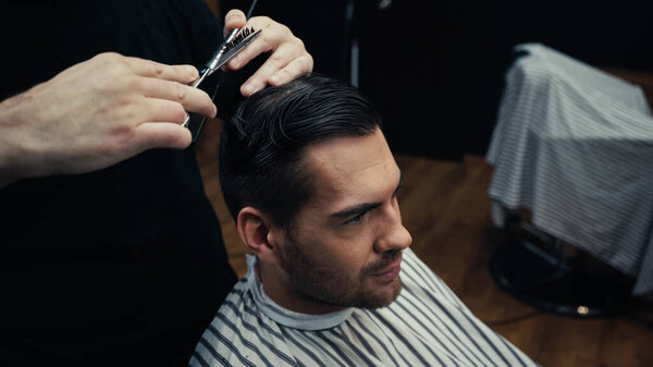 Hairstylist cutting hair of man in hairdressing cape in salon 