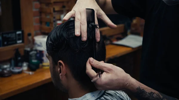 Barber combing hair of client and holding scissors in salon