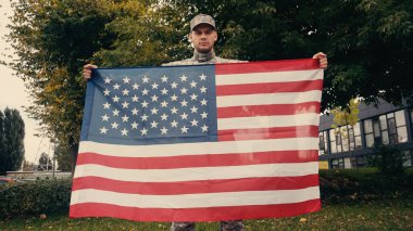 Askeri üniformalı ve şapkalı bir asker dışarıda kameralara bakarken Amerikan bayrağı taşıyor. 