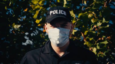 Ağacın yanında duran üniformalı ve tıbbi maskeli genç polis portresi. 