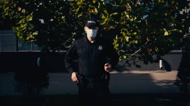 Dışarıda ağaçların yanında duran üniformalı ve sağlık maskeli bir polis memuru. 