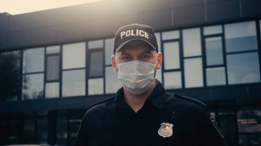 Tıbbi maskeli ve üniformalı bir polis memuru binanın yakınındaki kameraya bakıyor. 