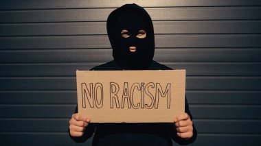 Kar maskeli bir adam elinde levhayla binanın yanında ırkçılık harfi yazmıyor. 