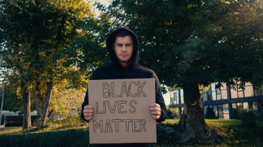 young activist in hoodie holding placard with black lives matter lettering outside  clipart