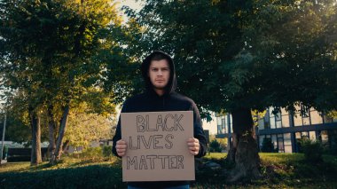 young man in hoodie holding placard with black lives matter lettering outside  clipart