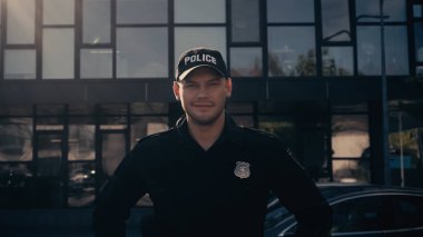 happy policeman looking at camera while standing in cap and uniform near building  clipart