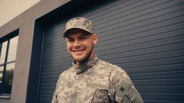 positive young soldier in uniform and cap smiling while looking at camera near building  clipart