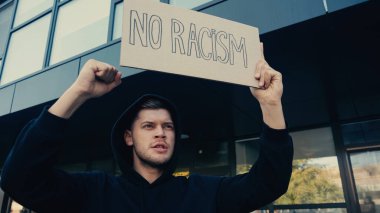 serious young man in hoodie holding placard with no racism lettering outdoors  clipart