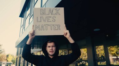 serious young activist in hoodie holding placard with black lives matter lettering on street clipart