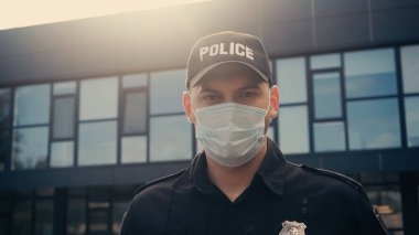 Maskeli ve üniformalı bir polis memuru şehir sokağındaki modern binanın yakınındaki kameraya bakıyor.