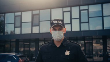 Tıbbi maskeli ve üniformalı bir polis memuru binanın yakınındaki kameraya bakıyor. 