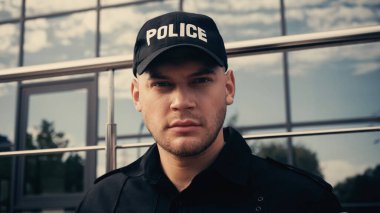 young policeman in cap with lettering police and uniform looking at camera clipart