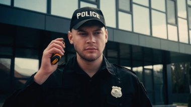 Üniformalı ve şapkalı genç bir polis telsizi açık havada tutarken bilgi dinliyor. 