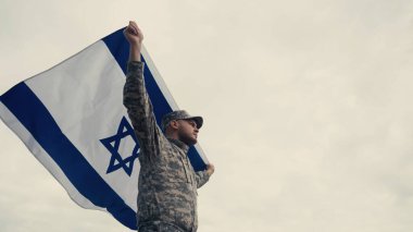 İsrail bayrağı taşıyan, arka planda gökyüzü olan üniformalı bir askerin alçak açılı görüntüsü. 